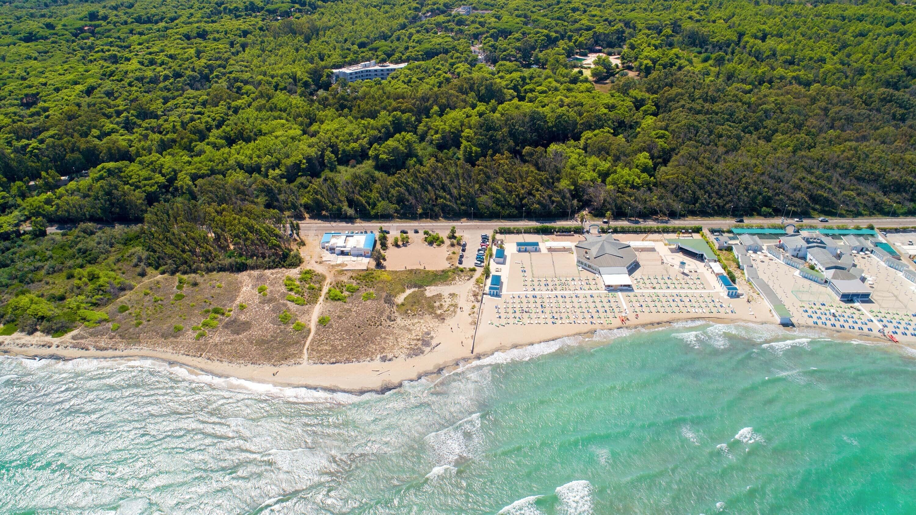 An aerial view of a coastal resort with a beach, a parking area, and a large building with multiple sections.