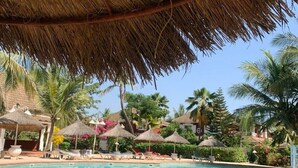 A pool area with thatched umbrellas and lounge chairs, surrounded by palm trees and a clear blue sky. - Dream villa KEUR BONHEUR in Saly: 3 suites, private pool, 100m from the beach (Saly)