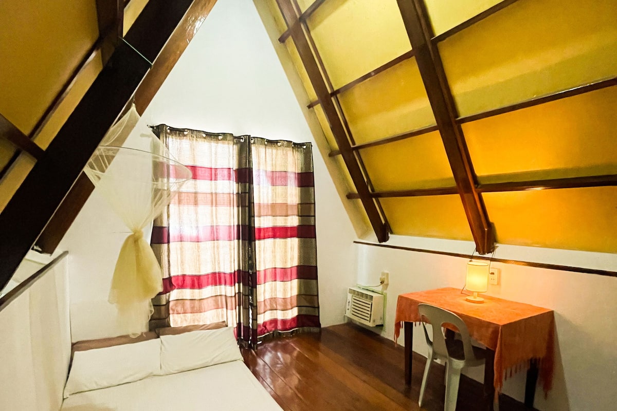 A room with a sloped ceiling, a bed, a table with a chair, and a window with curtains.