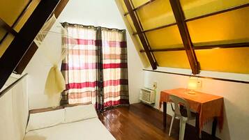 Una habitación con techo inclinado, una cama, una mesa con silla y un ventanal con cortinas.