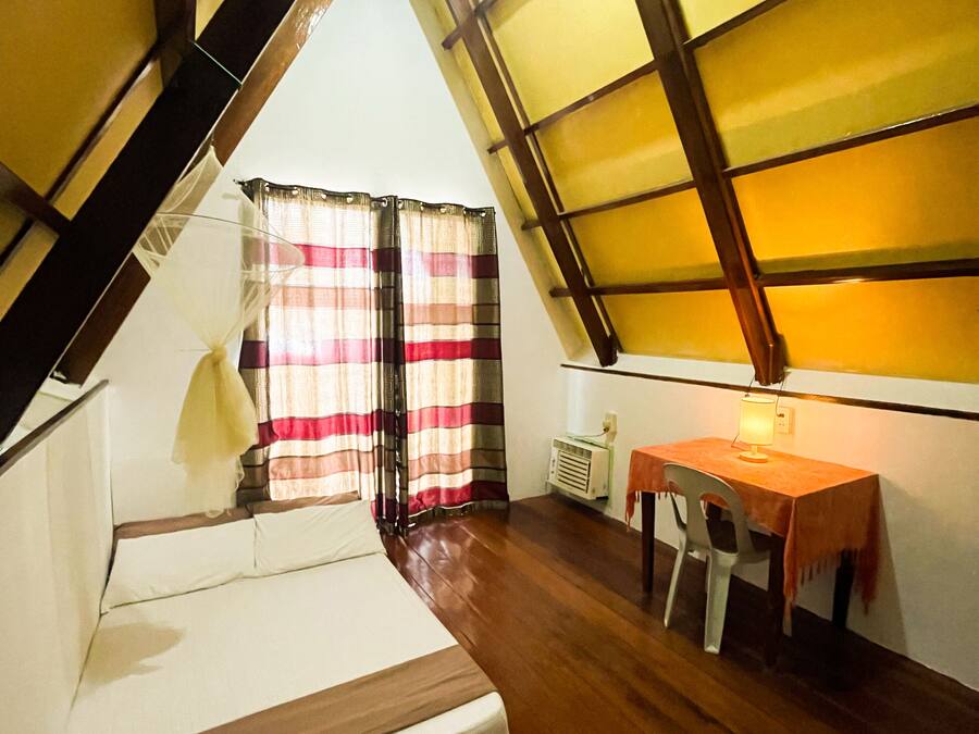 Una habitación con techo inclinado, una cama, una mesa con silla y un ventanal con cortinas.