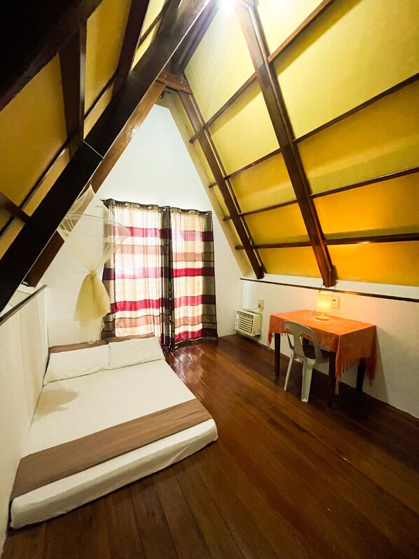 A room with a sloped ceiling, a bed, a table with a chair, and a window with curtains.