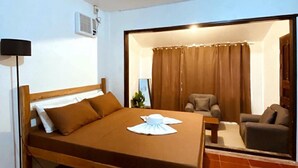 A single bed with a wooden frame, a bedside table, a lamp, and a window with curtains. - Flo Island Home (General Luna)