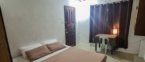 A hotel room with a bed, a desk, a chair, a lamp, and a door.