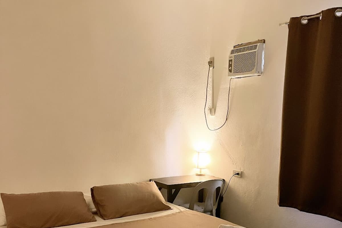 A hotel room with a bed, a small table with a lamp, a power outlet, and a window with curtains.