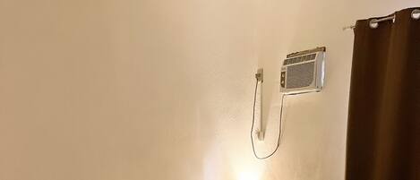 A hotel room with a bed, a small table with a lamp, a power outlet, and a window with curtains.