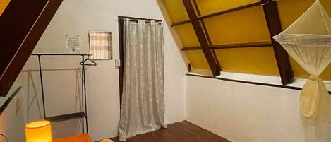 A small, cozy room with a wooden table, a bed, a window with a mosquito net, and a staircase leading to an upper level.
