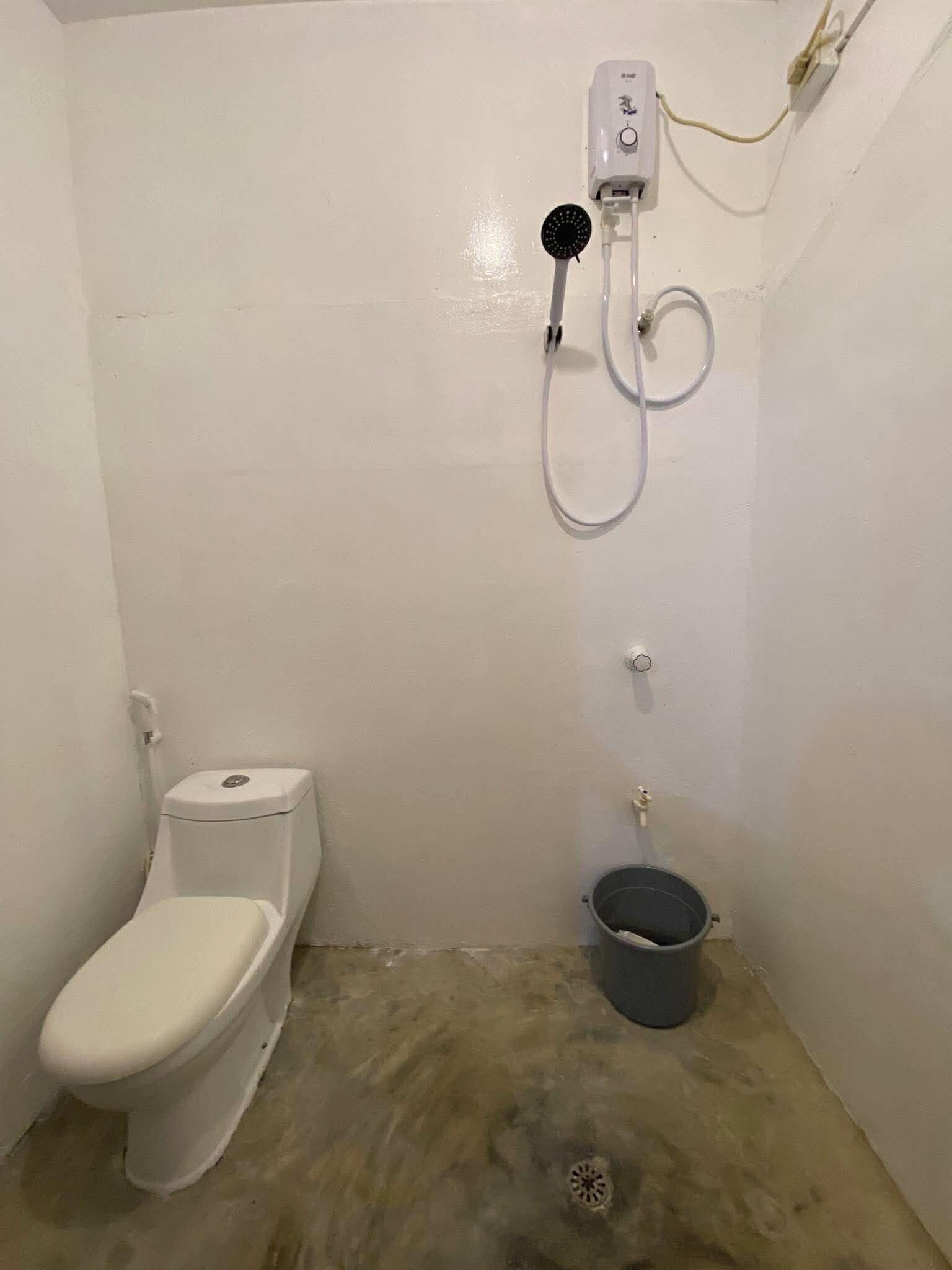 A small, enclosed bathroom with a toilet, a shower unit, and a bucket.
