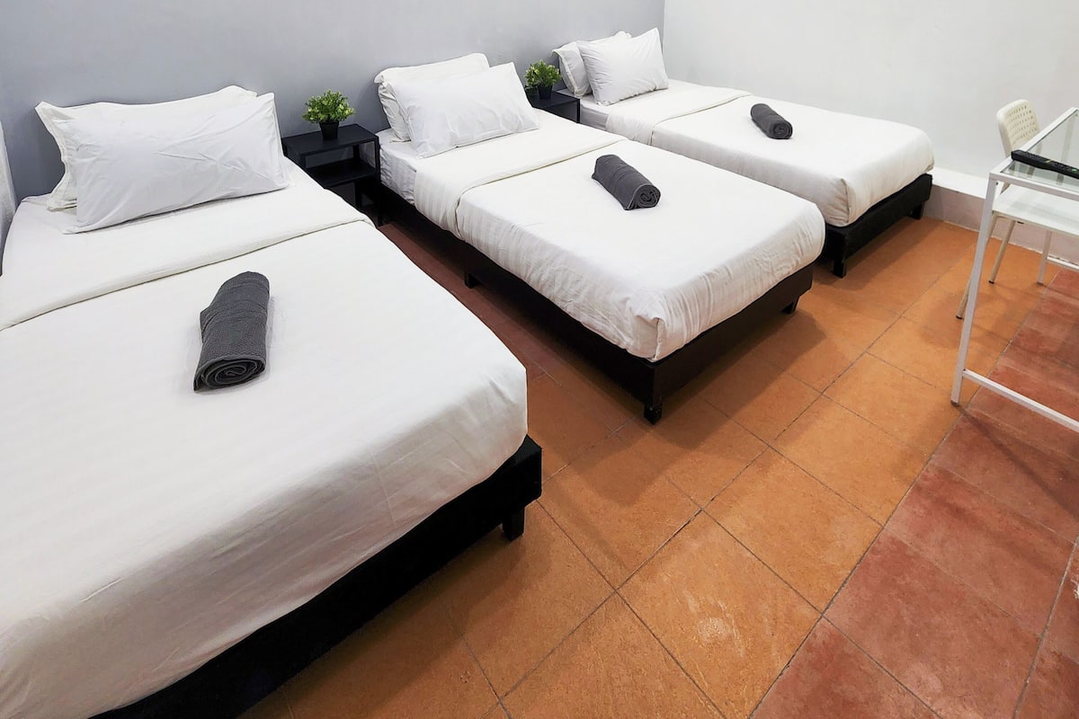 A hotel room with three single beds, each with white linens and a rolled towel. There is a small table with a remote control and a white chair.