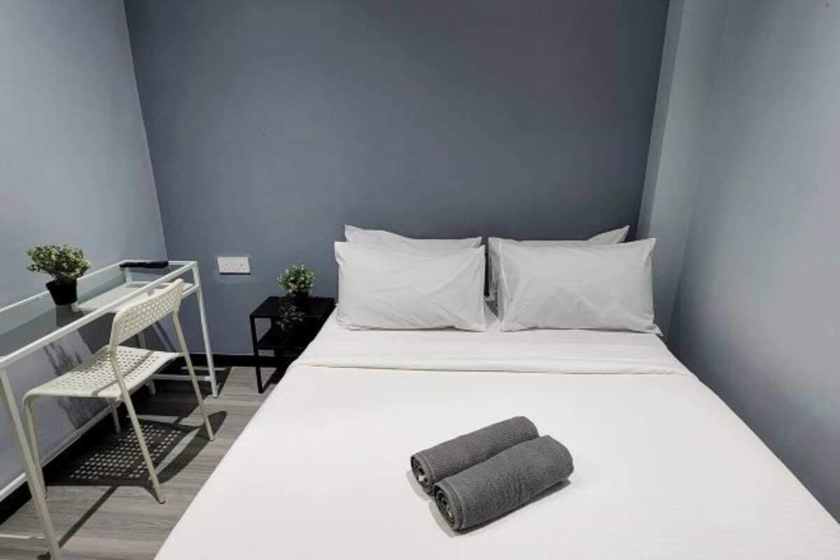 Habitación de hotel ordenada con una cama, dos almohadas, una toalla enrollada, una mesita con una planta y una silla.