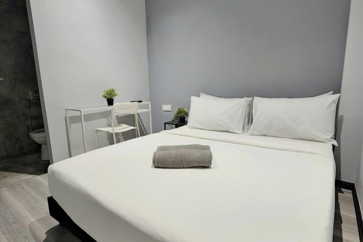 Un lit bien fait, avec du linge de lit blanc et une serviette pliée, un petit bureau blanc avec une chaise et une plante en pot.