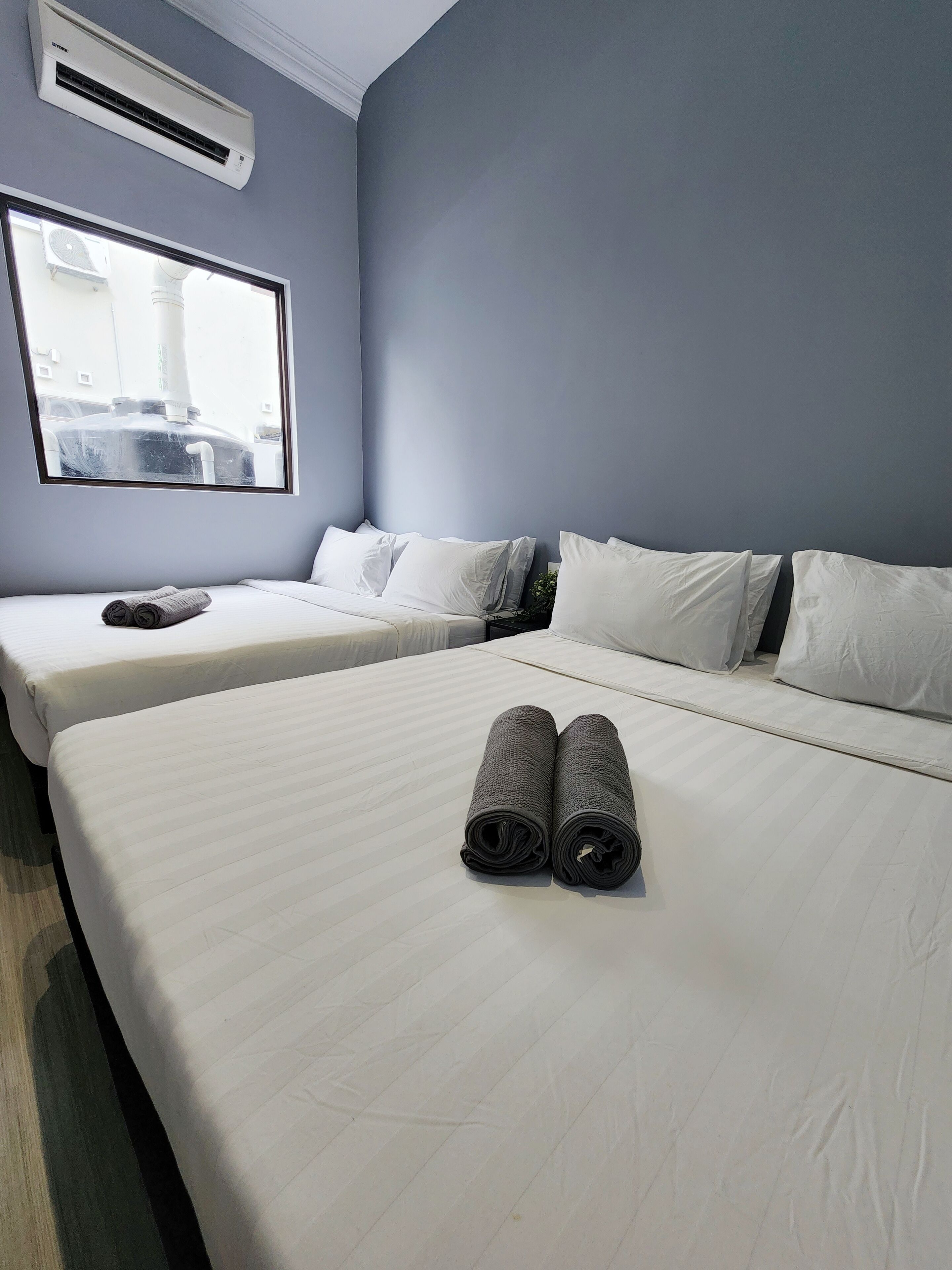 A hotel room with two beds, white bedding, a large window with a view of industrial structures, and an air conditioning unit on the wall.