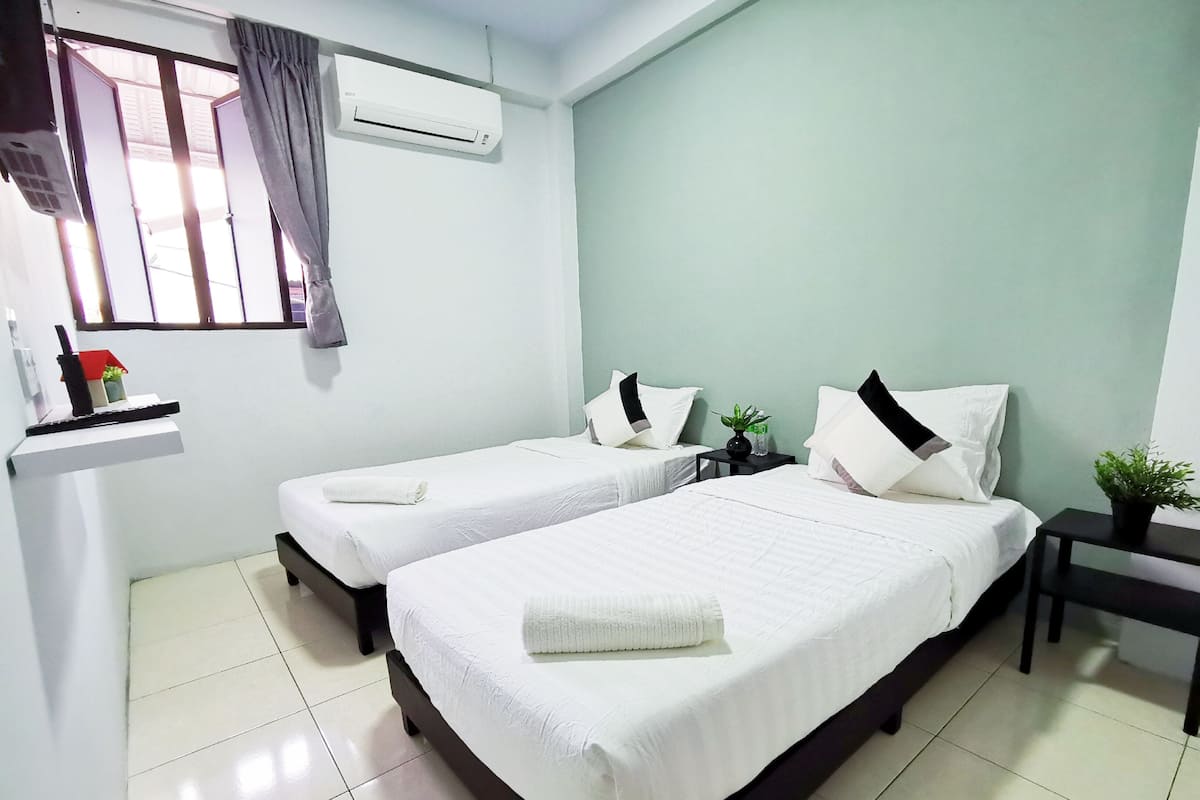 A hotel room with two single beds, a small table, a nightstand, an air conditioning unit, a window with curtains, and a shelf with a plant and a bottle.