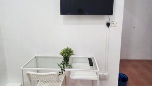 Una habitación pequeña con un escritorio blanco, una silla, una planta en maceta y un televisor montado.
