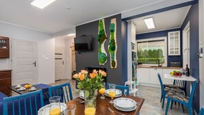Un área de comedor con una mesa lista para cuatro personas, un televisor montado en la pared y una cocina visible al fondo.