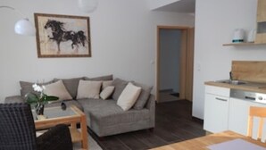 Una sala de estar con un sofá gris, una mesa de centro de madera y una mesa de comedor con individuales. Se observa una zona de cocina con lavavajillas y encimera de madera. En la pared, hay un cuadro enmarcado.