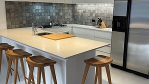 Una cocina moderna con una isla grande, taburetes de madera, un refrigerador de acero inoxidable y lámparas colgantes verdes.