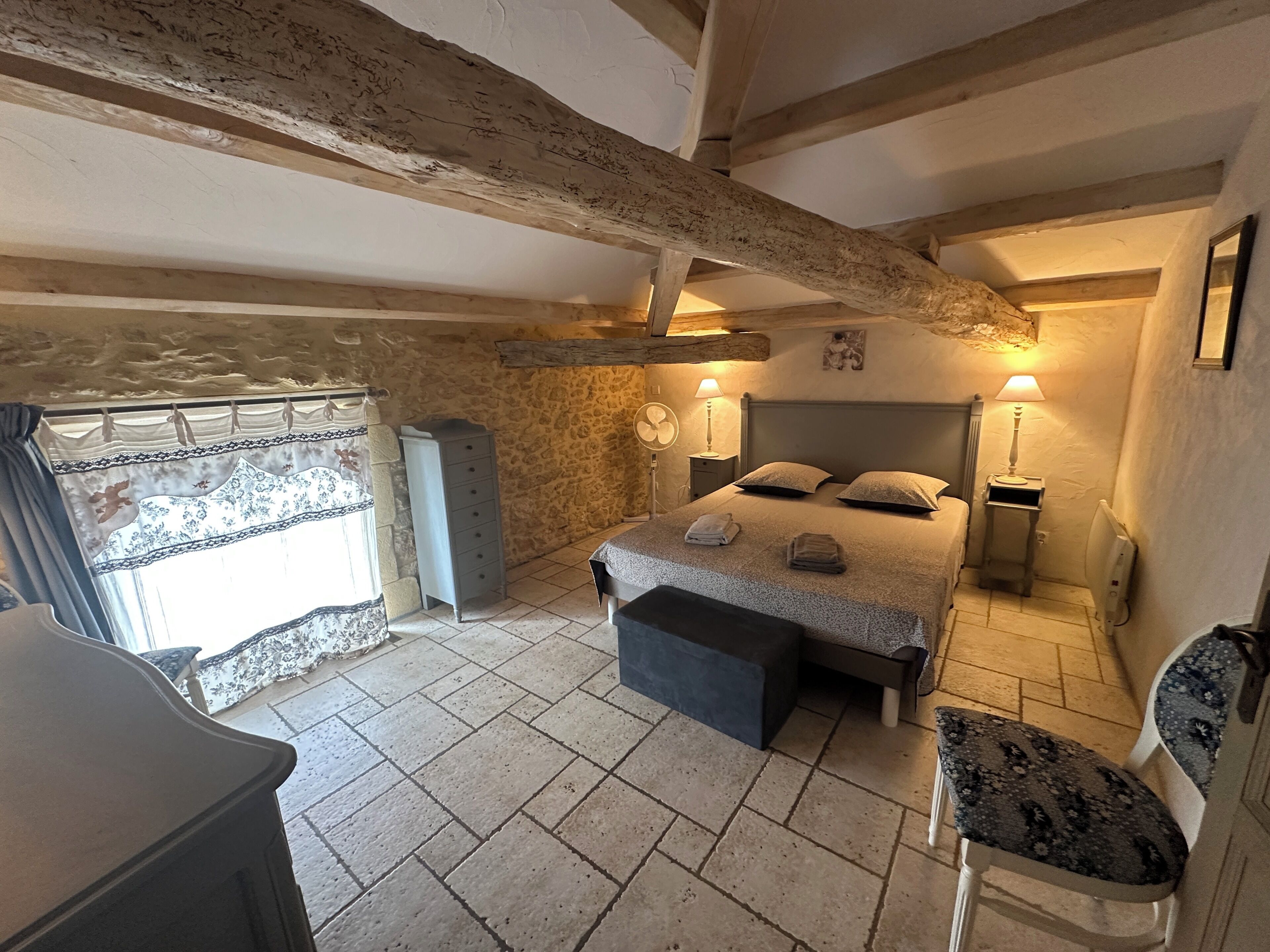 Une chambre à coucher avec un lit, des lampes de chevet, une commode, une chaise et une fenêtre avec des rideaux.