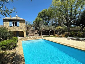 Une piscine aux murs de pierre, un bâtiment en pierre avec des volets bleus, et des arbres tout autour.