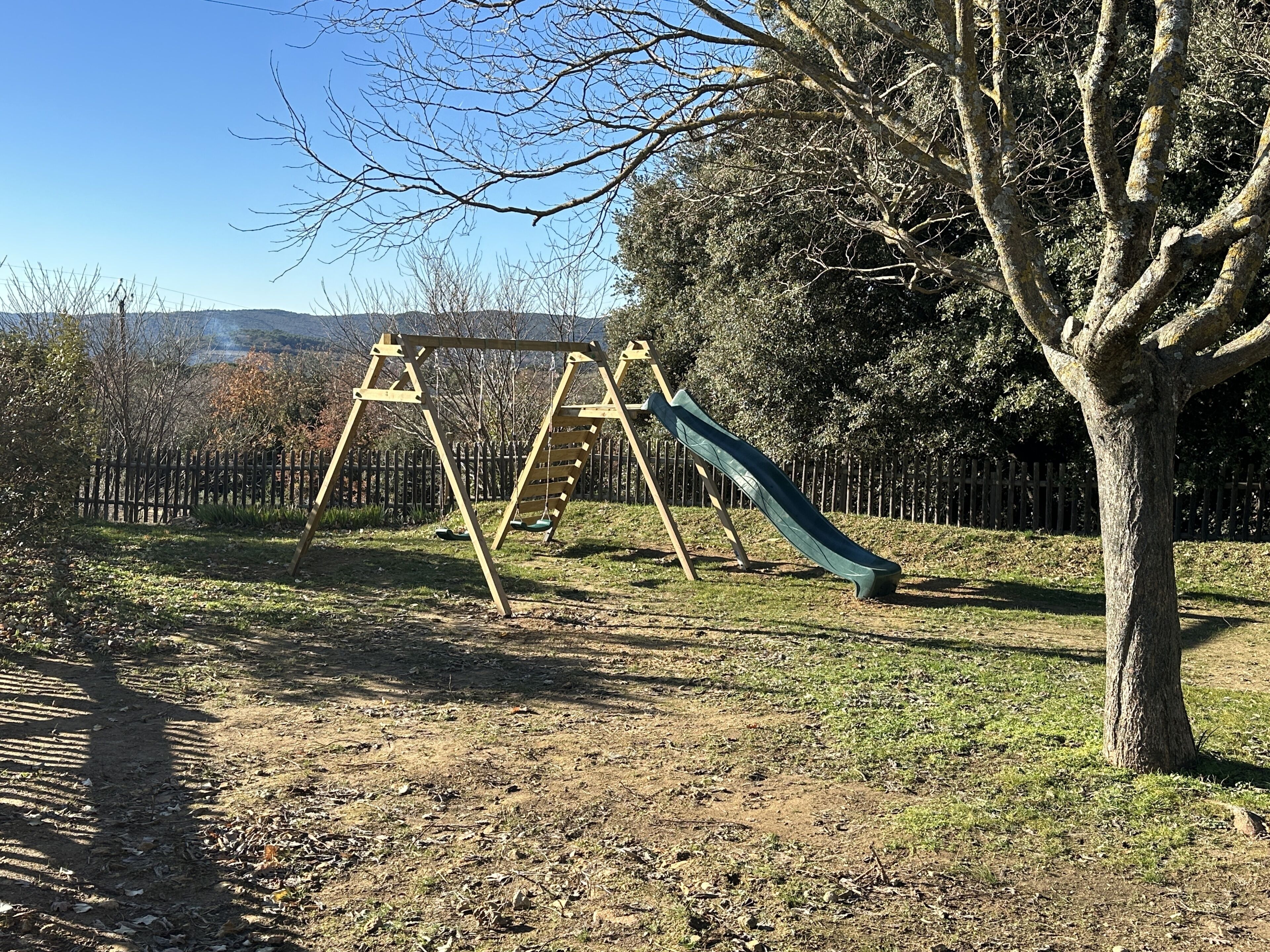 A playground with swings and a slide, surrounded by trees and a fence.