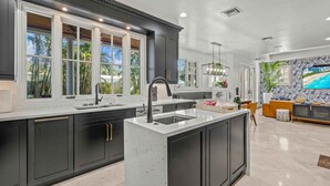 A modern kitchen with dark cabinetry, a central island, and large windows. - Walk to Beach / Heated Pool / Serenity at its Finest / Hemingway Key / VlLLAS (Fort Lauderdale)