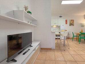 A modern living space with a flat-screen TV, white shelving, a dining area with green chairs, and a kitchen with white cabinets.