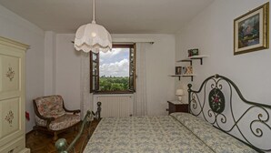 Ein Schlafzimmer mit Bett, Stuhl, Fenster mit Blick, Heizkörper, Regal mit Büchern und einem gerahmten Bild an der Wand.