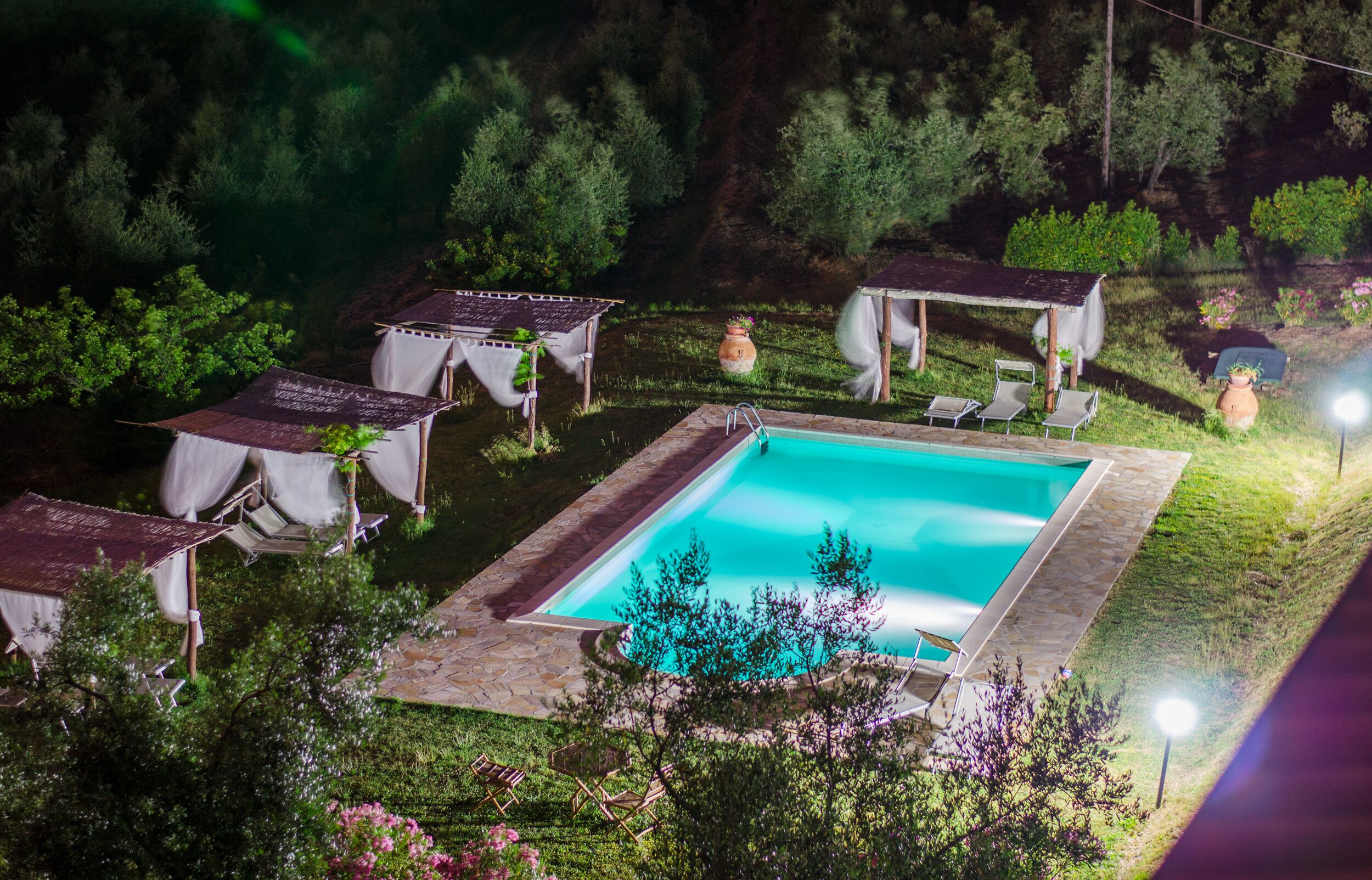 En swimmingpool omgiven av solstolar och möblerade sittgrupper, belägen i en frodig trädgård med träd och blommor.