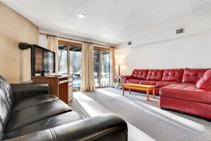Un salon avec un canapé rouge, un divan en cuir noir, une table basse en bois, un téléviseur et une porte-fenêtre coulissante donnant sur un balcon.