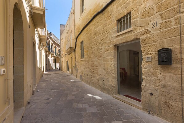 A narrow stone-paved street flanked by buildings with arched doorways and windows, one building has a number '21' displayed.