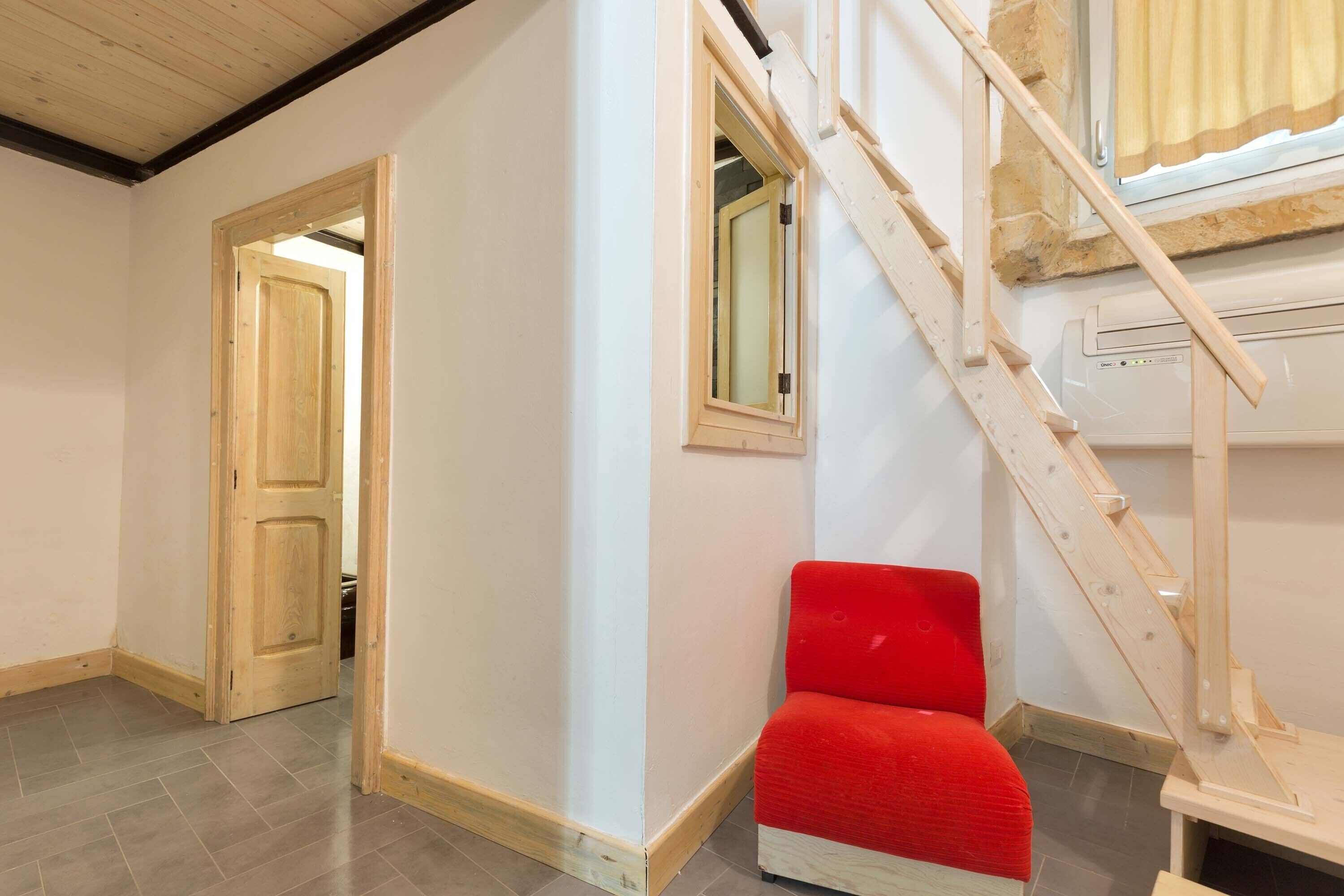 Una habitació amb una escala de fusta, una cadira vermella i una porta que dona a una altra habitació.
