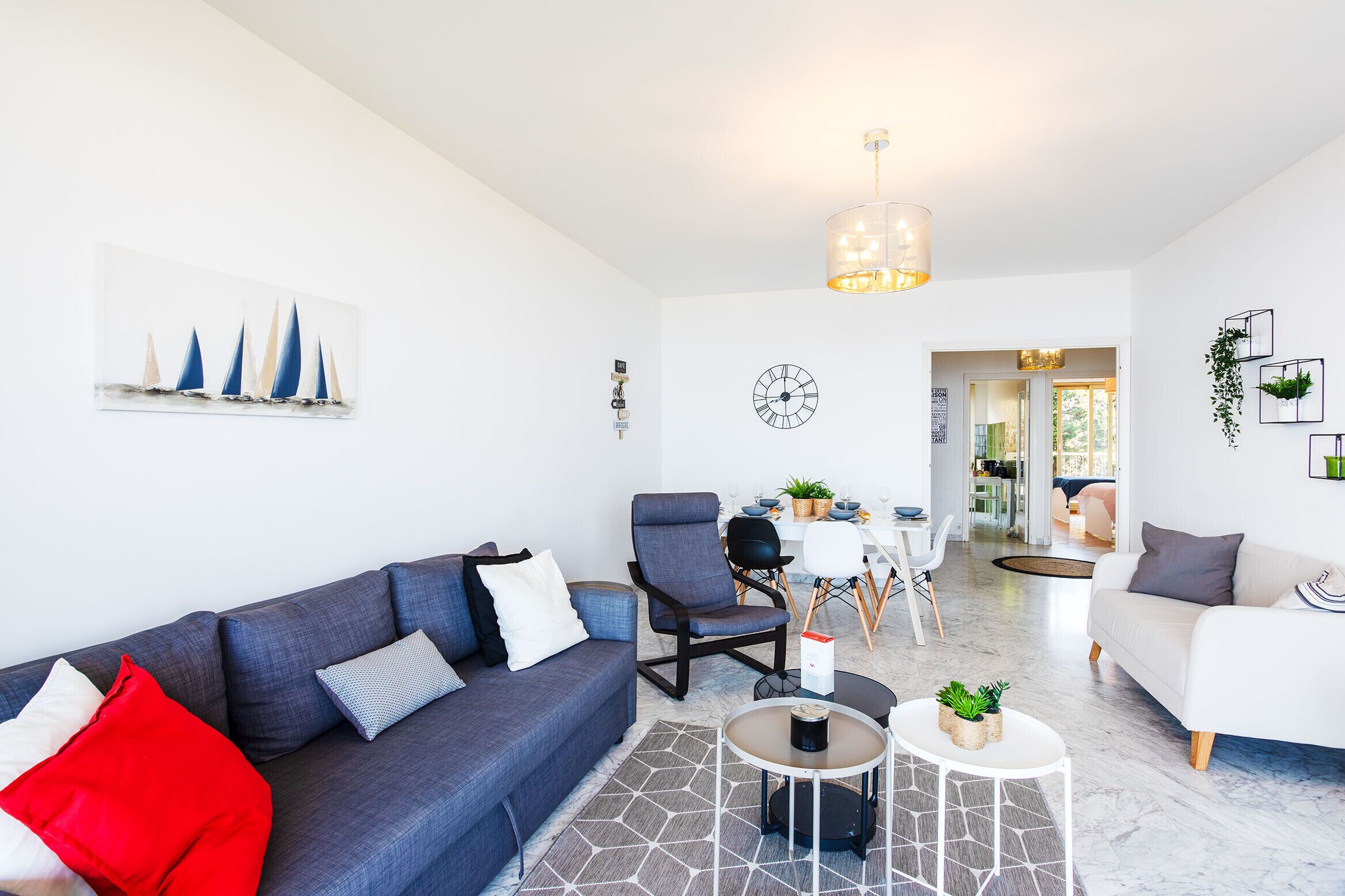 Un salon moderne avec un canapé gris, un canapé blanc et un coin repas comprenant une table et des chaises. Il y a une horloge au mur et une plante sur le côté.