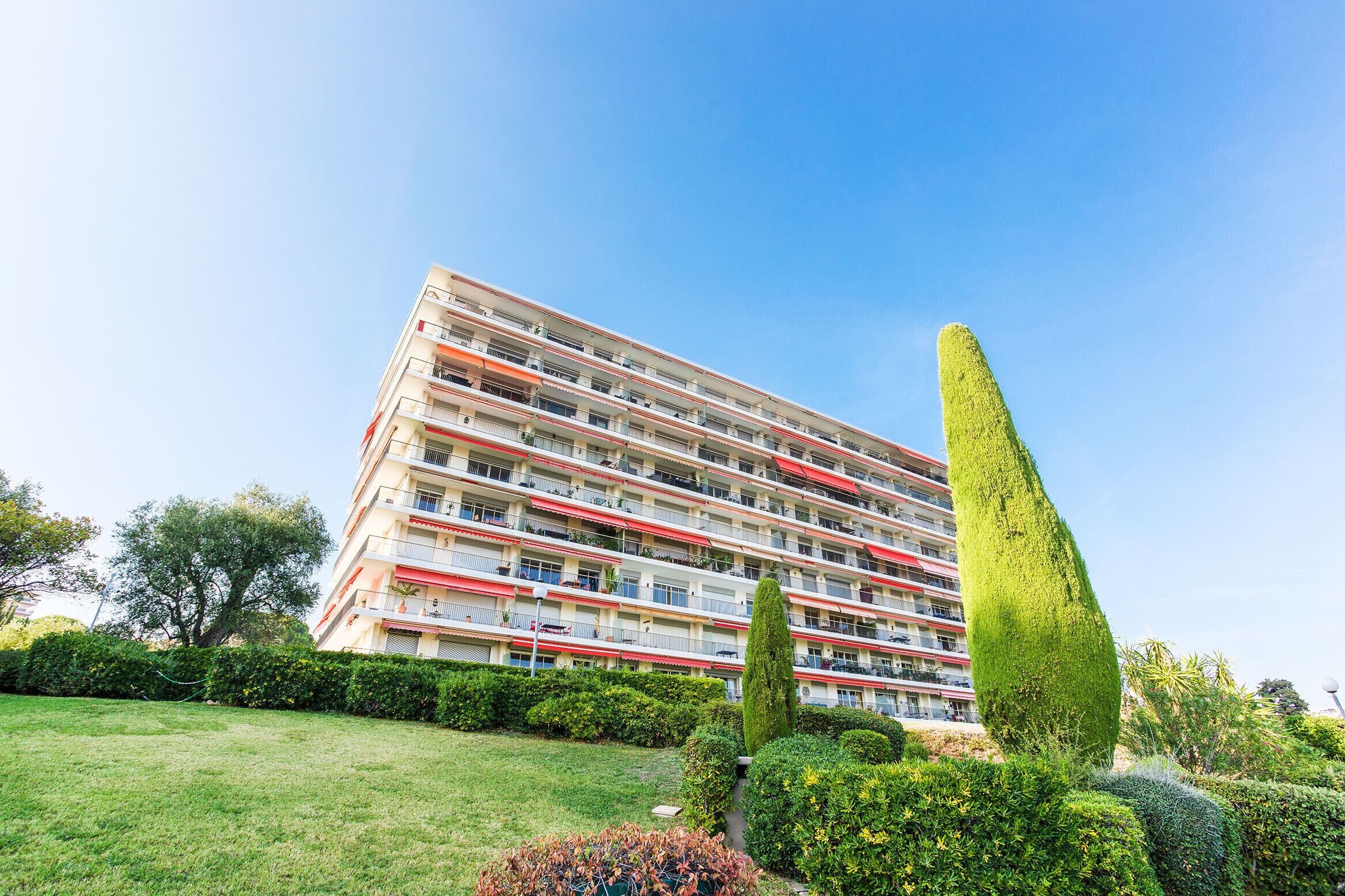 A multi-story residential building with balconies, surrounded by well-maintained gardens and hedges.