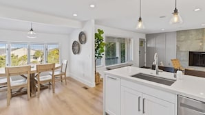 A modern kitchen with white cabinetry, a large island with a sink, pendant lighting, and a view of the outdoors through large windows. - @ Marbella Lane - 3BR w/sweeping views of Bay Area (San Mateo)