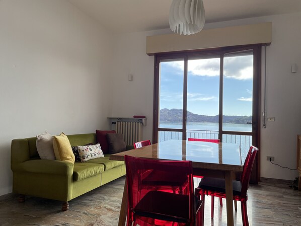 A living room with a green sofa, red chairs, a glass-top dining table, and a large window offering a view of a lake and hills.