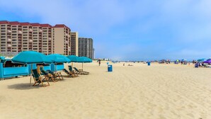 Une plage avec des parasols bleus et des chaises longues, un rivage sablonneux et, en arrière-plan, des immeubles de grande hauteur.