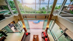 A spacious indoor area with a large window overlooking a pool and lush greenery. - Glasshouse Phuket | 6BR Villa | Sleeps 22 | Walk to 2 Beaches | Accessible (Vichit, Muang)