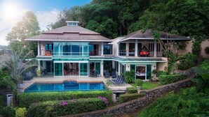 A two-story house with a large glass facade, a swimming pool, and lush greenery surrounding the property. - Glasshouse Phuket | 6BR Villa | Sleeps 22 | Walk to 2 Beaches | Accessible (Vichit, Muang)