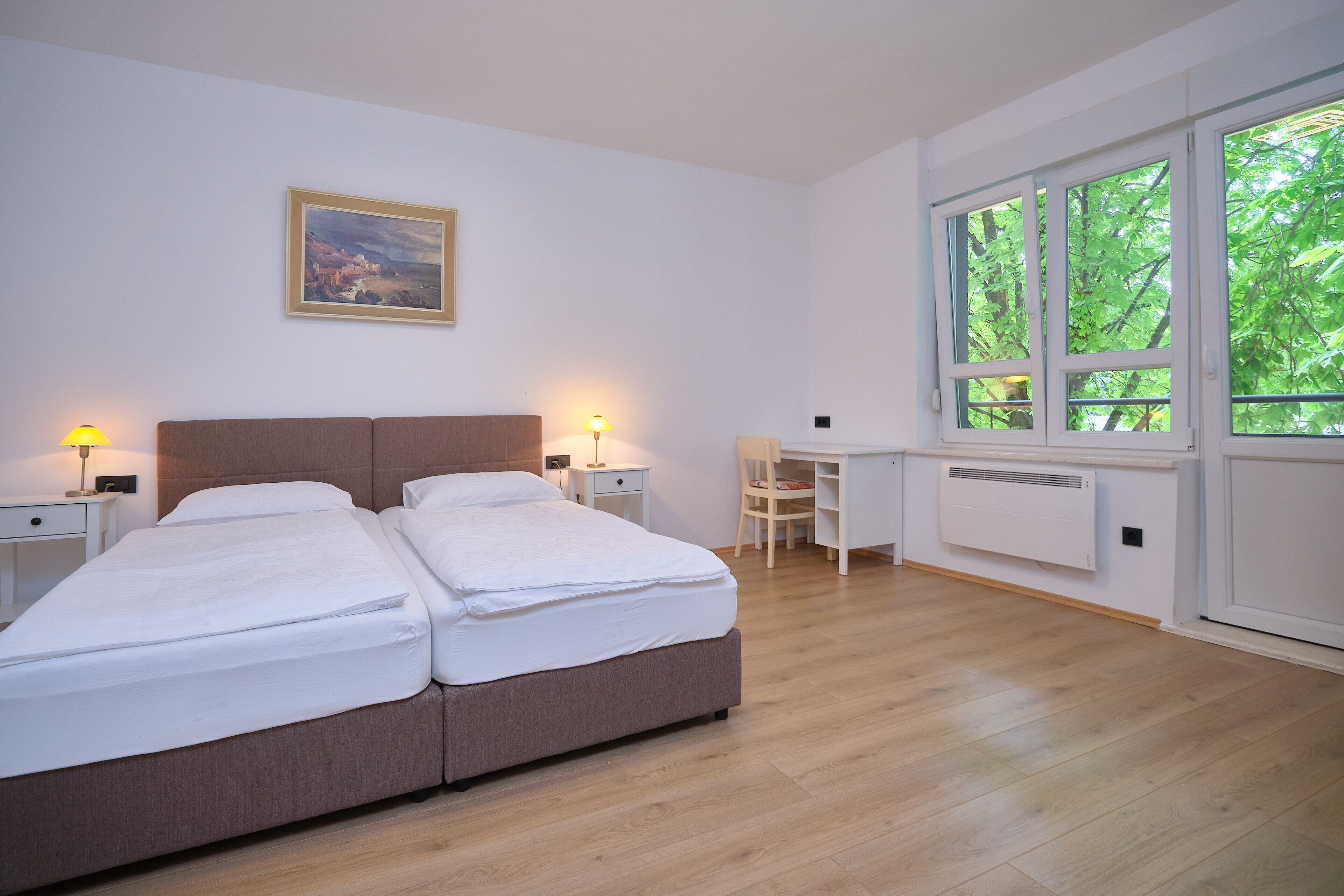 A bedroom with a double bed, bedside tables with lamps, a desk, and a window with a view of trees.