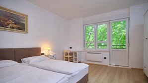 Camera d'albergo con due letti, una scrivania, una sedia, un termosifone e un'ampia finestra con vista su un'area verde.