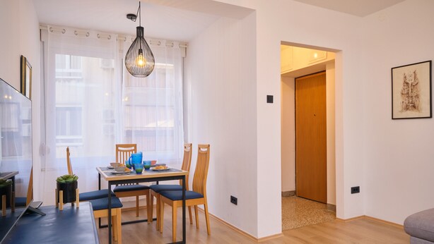 Un soggiorno moderno con zona pranzo, pavimento in legno, un'ampia finestra con tende leggere e una porta che dà su un'altra stanza.