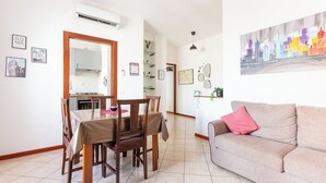 A dining area with a wooden table and chairs, a kitchen in the background, and a living room with a sofa and a cityscape painting. - Camesena 40 | 2BR Apartment for 4 with Balcony near Tiburtina (Roma)