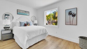 Une chambre à coucher avec un lit, des tables de chevet, une fenêtre donnant sur des palmiers et deux tableaux encadrés accrochés au mur.
