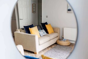 A cozy living room with a beige sofa, yellow and blue cushions, a small round ottoman, and a white radiator.