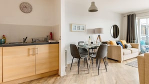 A modern kitchen with wooden cabinets, a dining area with a table and chairs, a sofa, and a large window with curtains.