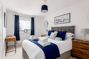 A neatly arranged bedroom with a bed, bedside tables, a window with curtains, a wall clock, and a framed picture.