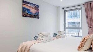 A neatly made bed with a pink blanket and pillows, a stack of folded towels, and a painting of a coastal landscape on the wall. - Once Upon a Tide - Apartment in the Heart of Poole (Poole)