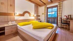 A bedroom with a wooden ceiling, a bed with yellow pillows, a nightstand, a desk, and a window with a view of trees. - Holiday Apartment "Niederkoflhof Wollbach" with Mountain View, Balcony & Wi-Fi (Valle Aurina)