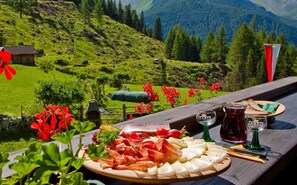 Ein malerischer Bergblick mit einem Holztisch, auf dem eine Platte mit Wurst- und Käsesorten, ein Glas Rotwein und eine Marmeladenglas stehen.