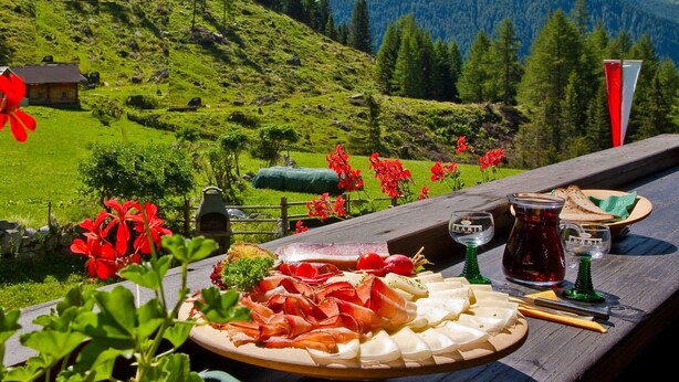 A scenic mountain view with a wooden table displaying a charcuterie board, a glass of red wine, and a bottle of jam.